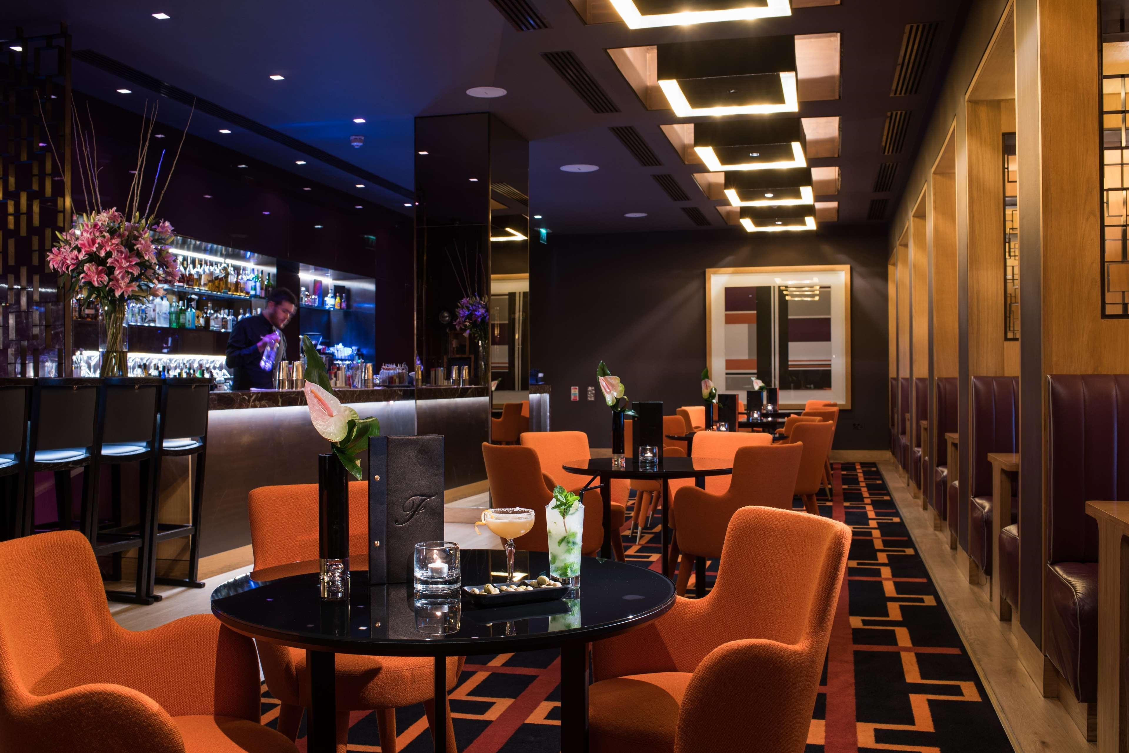 The Fitzwilliam Hotel Белфаст Екстериор снимка The photo depicts a stylish bar or lounge area with a modern and sophisticated atmosphere. The space features dark walls accented by blue lighting and a sleek bar counter in the background, stocked with various drinks. The seating consists of round t