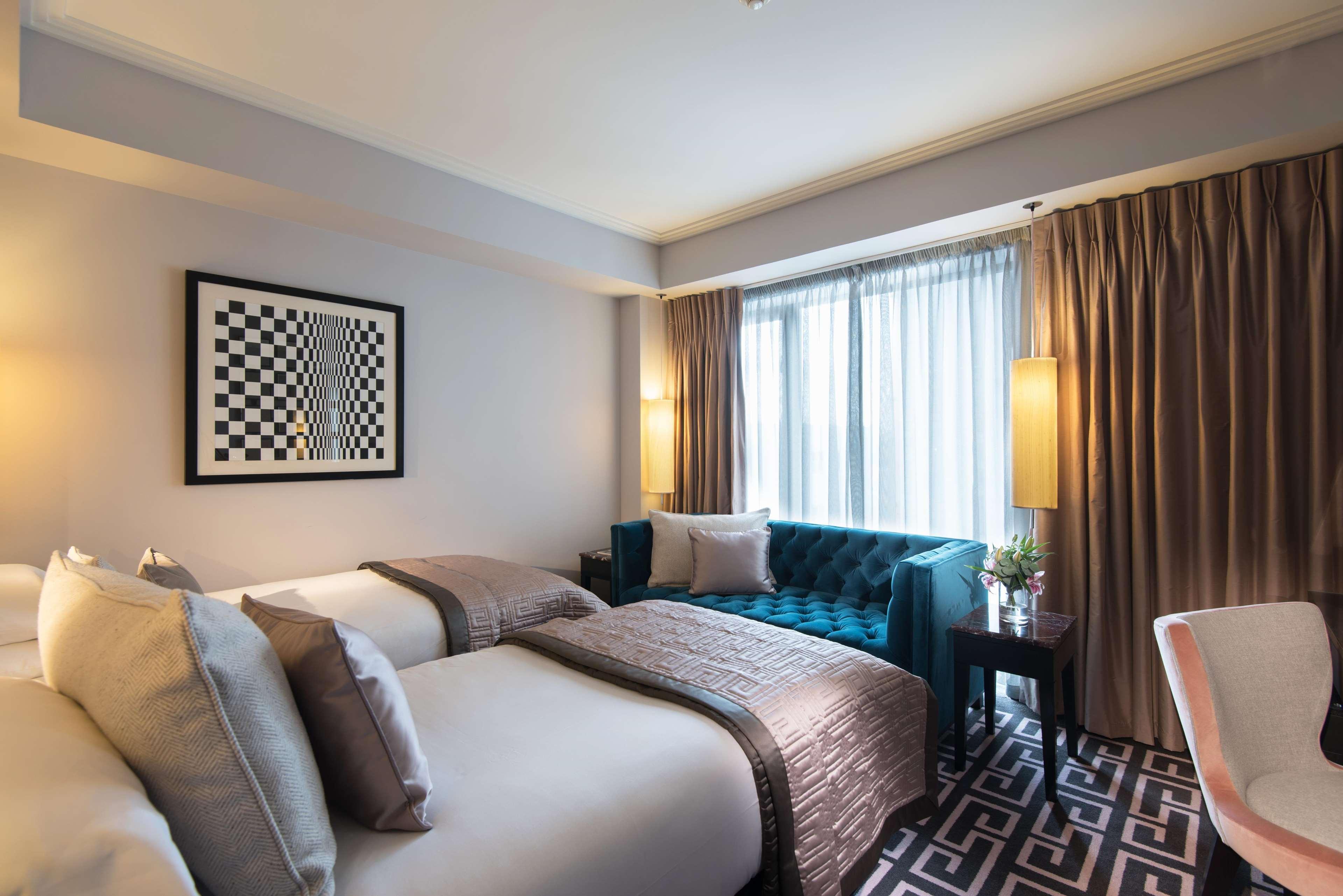 The Fitzwilliam Hotel Белфаст Екстериор снимка The photo shows a modern hotel room. It features two beds with decorative pillows, a stylish teal couch, and large windows with sheer curtains allowing natural light to enter. The walls are painted in neutral tones, and there is a piece of artwork wi