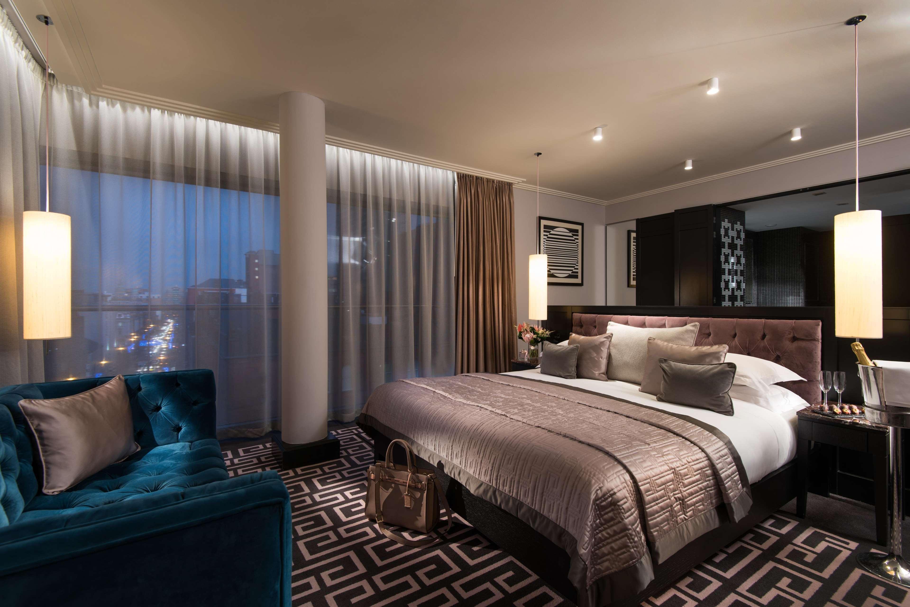 The Fitzwilliam Hotel Белфаст Екстериор снимка The photo shows a luxurious hotel room with a modern design. It features a large bed with decorative pillows and a stylish bedspread, complemented by bedside lamps. To one side, there is a plush turquoise sofa and a coffee table. The room has floor-t