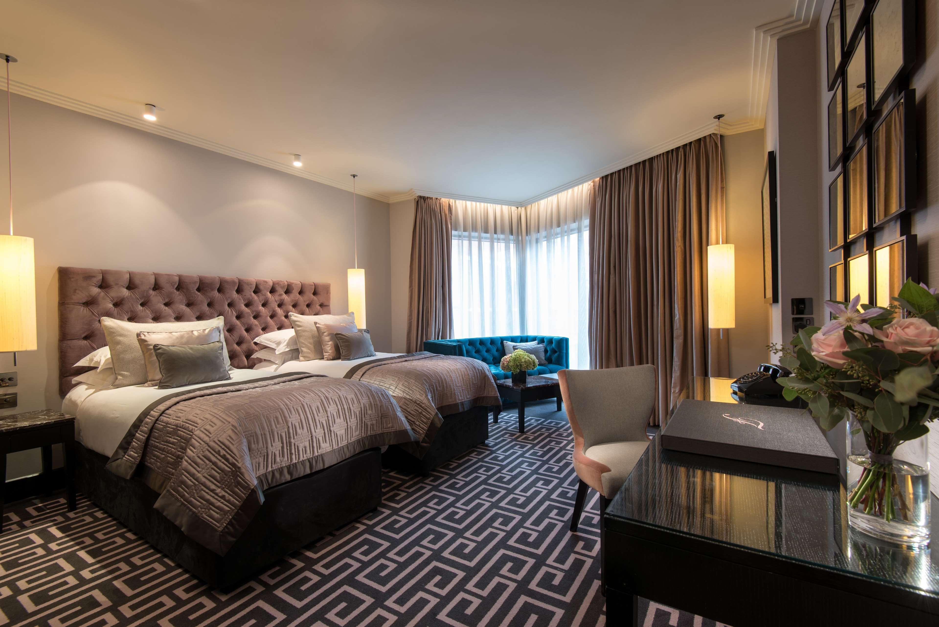 The Fitzwilliam Hotel Белфаст Екстериор снимка The photo shows a stylish hotel room featuring two twin beds with tufted headboards and elegant bedding. On one side, there is a luxurious blue couch. Large windows are covered with light curtains, allowing natural light to enter. The floor is adorne
